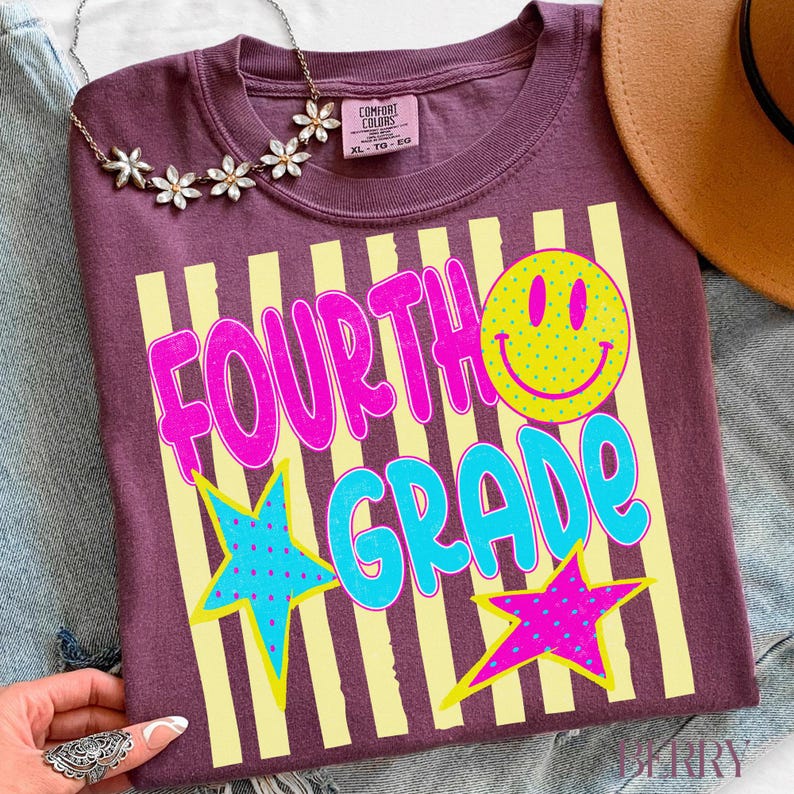 Op de afbeelding: Een bordeauxrood T-shirt met de woorden "FOURTH GRADE" in felroze en blauw, een gele smiley en twee sterren. Het shirt heeft gele verticale strepen en een Comfort Colors label.