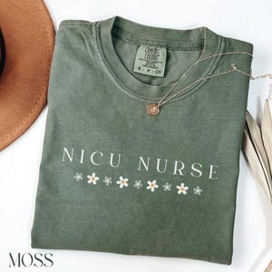 Peut inclure: Un t-shirt vert avec le texte "NICU NURSE" en lettres blanches et une rang&eacute;e de marguerites blanches sous le texte. Le t-shirt est pli&eacute; et porte une &eacute;tiquette indiquant "COMFORT COLORS".