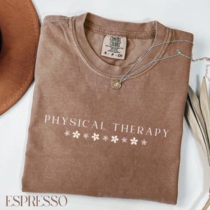 May include: Espresso-colored t-shirt with the words "PHYSICAL THERAPY" in white, with small daisy graphics underneath. A silver necklace with a pendant is draped across the shirt. The shirt is a Comfort Colors brand.