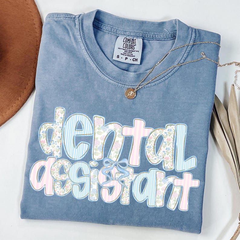 Op de afbeelding: Een blauw Comfort Colors T-shirt met de woorden "dental assistant" in een bloemen- en streepjesontwerp. Een gouden ketting met een hanger is over het shirt gedrapeerd. Het shirt is opgevouwen en heeft een klein label.