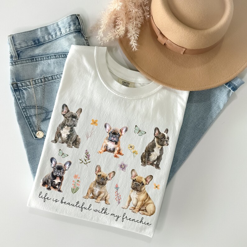 May include: White t-shirt featuring a graphic of six French Bulldogs and flowers. The text "life is beautiful with my frenchie" is printed on the shirt.