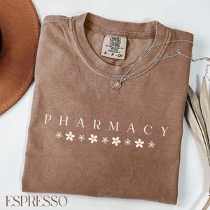 May include: A brown t-shirt with the word "PHARMACY" printed in white with a row of white daisies underneath. The shirt has a tag that says "COMFORT COLORS" and "S - P - CH".