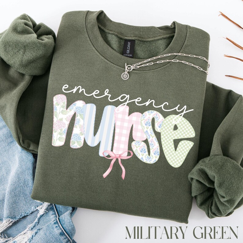 Puede incluir: Sudadera verde militar con la frase "emergency nurse". La palabra "nurse" est&aacute; decorada con estampados florales, cuadros y rayas. Un collar plateado con un peque&ntilde;o colgante est&aacute; en la sudadera.