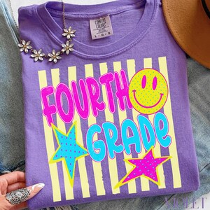 Op de afbeelding: Een paars Comfort Colors T-shirt met de woorden "FOURTH GRADE" in roze en blauw, samen met een gele smiley en sterren. Het shirt heeft een ronde hals en korte mouwen. Maat L-G.