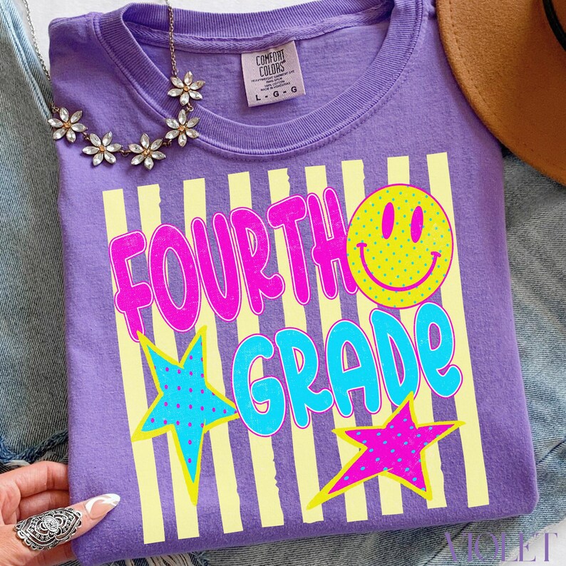 Op de afbeelding: Een paars Comfort Colors T-shirt met de woorden "FOURTH GRADE" in roze en blauw, samen met een gele smiley en sterren. Het shirt heeft een ronde hals en korte mouwen. Maat L-G.