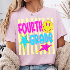 Op de afbeelding: Lichtroze T-shirt met de tekst "FOURTH GRADE" in felroze en blauw, met een gele smiley en sterren. Het ontwerp heeft gele verticale strepen. Een vrolijk ontwerp, perfect voor het nieuwe schooljaar.