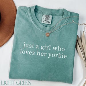 May include: Light green t-shirt with the text "just a girl who loves her yorkie" in white. A silver necklace with a round pendant rests on the shirt. The shirt is folded and has a Comfort Colors tag.
