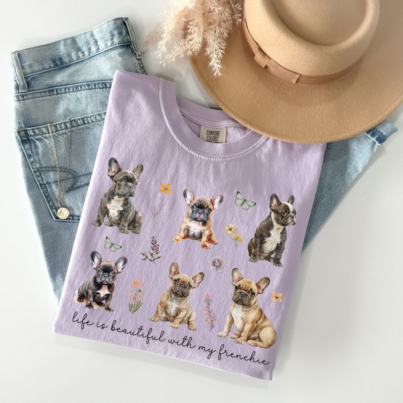May include: A lavender t-shirt featuring a graphic print of six French Bulldogs and the text "Life is beautiful with my frenchie".