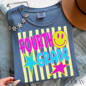 Op de afbeelding: Een blauw t-shirt met de woorden "FOURTH GRADE" in roze en blauw, een gele smiley en sterren. Het shirt heeft gele verticale strepen en is van het merk Comfort Colors.