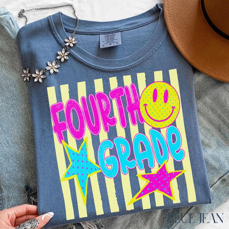Op de afbeelding: Een blauw t-shirt met de woorden "FOURTH GRADE" in roze en blauw, een gele smiley en sterren. Het shirt heeft gele verticale strepen en is van het merk Comfort Colors.