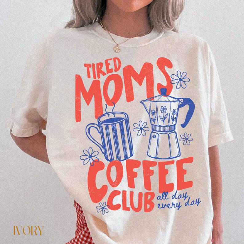 Puede incluir: Camiseta de color crema con la frase "Tired Moms Coffee Club" en rojo y azul. El dise&ntilde;o incluye una taza de caf&eacute;, una cafetera y motivos florales. Tambi&eacute;n se incluye la frase "all day every day".