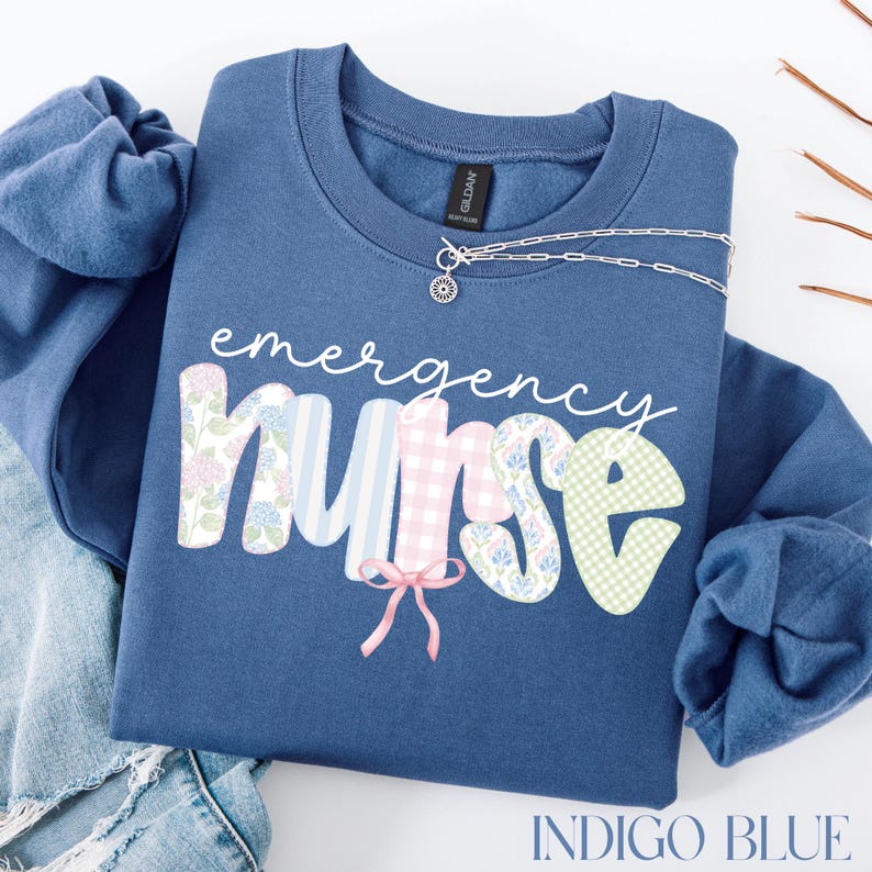 Puede incluir: Sudadera azul &iacute;ndigo con la frase "emergency nurse" en una fuente decorativa. Las letras est&aacute;n estampadas con dise&ntilde;os florales, de cuadros y a rayas. Un collar plateado con un colgante de sol est&aacute; drapeado en la parte superior.