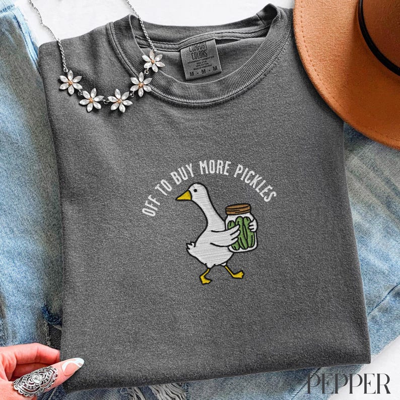 May include: Dark gray Comfort Colors t-shirt with the phrase "OFF TO BUY MORE PICKLES" and a cartoon goose carrying a jar of pickles. The shirt has a crew neck and short sleeves. A silver necklace with flower accents is visible.