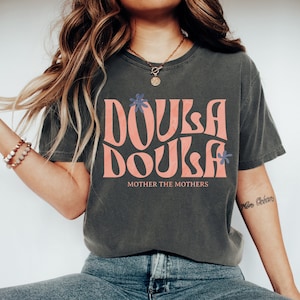 May include: A dark gray t-shirt with a pink graphic that says "Doula Doula" with blue flowers. The text "Mother the Mothers" is printed below the graphic.