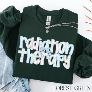 May include: Forest green sweatshirt with the words "radiation therapy" in a floral and striped design. A silver necklace with a sunburst pendant is on the sweatshirt. The sweatshirt is folded with a pair of blue jean shorts.