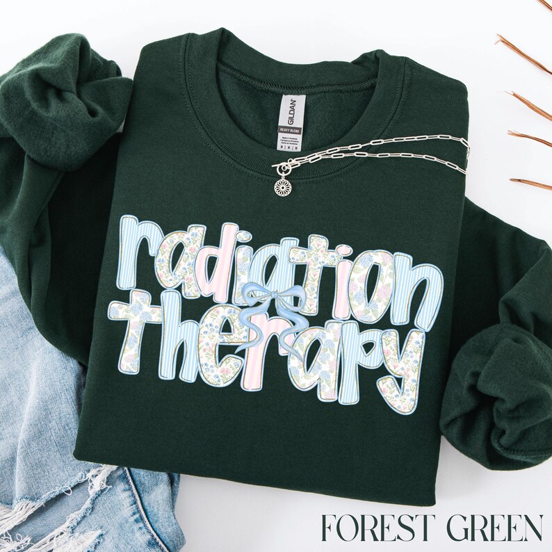 May include: Forest green sweatshirt with the words "radiation therapy" in a floral and striped design. A silver necklace with a sunburst pendant is on the sweatshirt. The sweatshirt is folded with a pair of blue jean shorts.