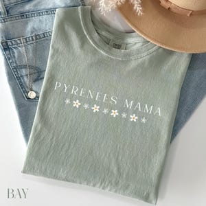 Puede incluir: Una camiseta verde claro con el texto "Pyrenees Mama" en letras blancas y una fila de margaritas blancas con centros amarillos.