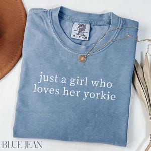 May include: Blue jean colored t-shirt with the text "just a girl who loves her yorkie" in white. A gold necklace with a pendant rests on the shirt. The shirt has a Comfort Colors tag.