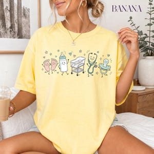 May include: A pale yellow short-sleeve t-shirt featuring a graphic print of cartoon baby items including footprints, a bottle, a bassinet, a stethoscope, and a pacifier, all with smiling faces, surrounded by small stars and hearts.