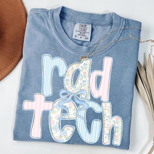 May include: A blue Comfort Colors t-shirt with the words "rad tech" in a whimsical design. The letters are decorated with floral patterns, stripes, and a light blue bow. A silver necklace is draped across the shirt.