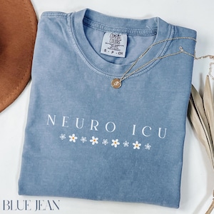 May include: A blue jean colored t-shirt with the words "NEURO ICU" in white, along with a row of white daisies. A gold necklace with a small pendant rests on the shirt. The shirt is folded and has a tag that says "Comfort Colors".