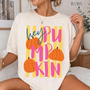 Peut inclure: Un t-shirt à manches courtes ivoire avec une impression graphique portant le texte "hey PUMPKIN" en bleu et rose. Le motif comprend trois citrouilles orange et des rayures verticales jaunes sur le devant du vêtement.