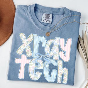 May include: A blue Comfort Colors t-shirt with the words "Xray Tech" in a floral and striped design. The shirt has a small bow and a gold necklace. The shirt is folded and has a tag that says "Comfort Colors".