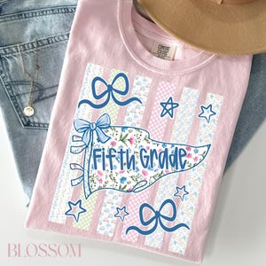 May include: A light pink t-shirt featuring a central pennant graphic with the text Fifth Grade. The design includes floral patterns, blue bows, and star shapes layered over vertical stripes. The shirt has a Comfort Colors brand neck label.