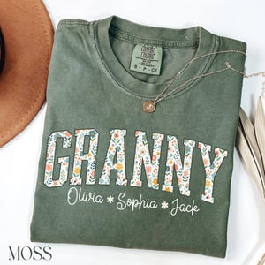May include: Green t-shirt with the word "Granny" in white letters with a floral pattern. The names "Olivia", "Sophia", and "Jack" are written below the word "Granny".