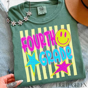 Op de afbeelding: Lichtgroen t-shirt met de woorden "FOURTH GRADE" in felroze en blauw, een gele smiley en twee sterrenafbeeldingen. Het shirt heeft een ronde hals en korte mouwen. Een bloemenketting en een bruine hoed zijn zichtbaar.