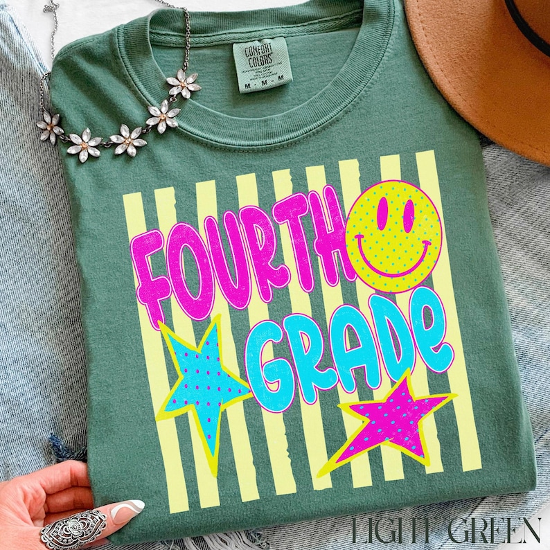 Op de afbeelding: Lichtgroen t-shirt met de woorden "FOURTH GRADE" in felroze en blauw, een gele smiley en twee sterrenafbeeldingen. Het shirt heeft een ronde hals en korte mouwen. Een bloemenketting en een bruine hoed zijn zichtbaar.