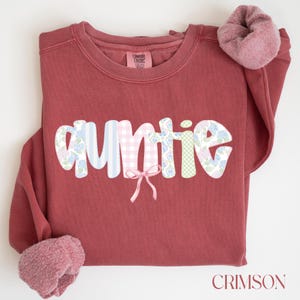 May include: Crimson-colored sweatshirt with the word "auntie" in a patchwork design. The letters feature floral, striped, and gingham patterns. A small pink bow is centered below the word. A matching scrunchie rests on the sleeve. The word "CRIMSON" is at the bottom.