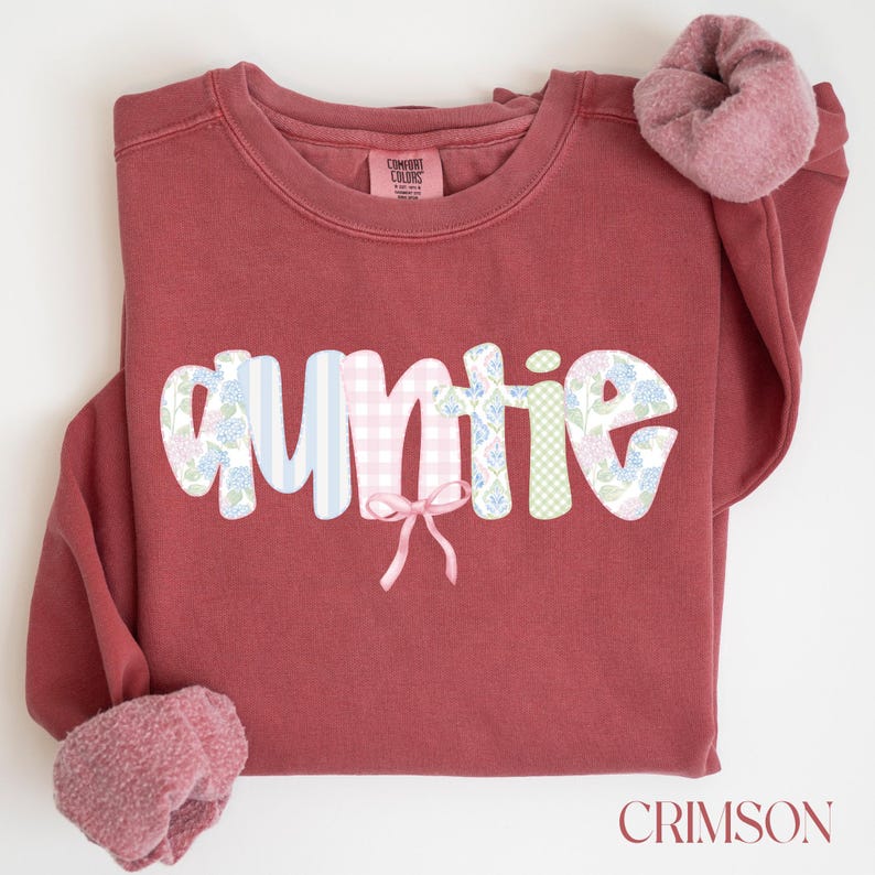 May include: Crimson-colored sweatshirt with the word "auntie" in a patchwork design. The letters feature floral, striped, and gingham patterns. A small pink bow is centered below the word. A matching scrunchie rests on the sleeve. The word "CRIMSON" is at the bottom.