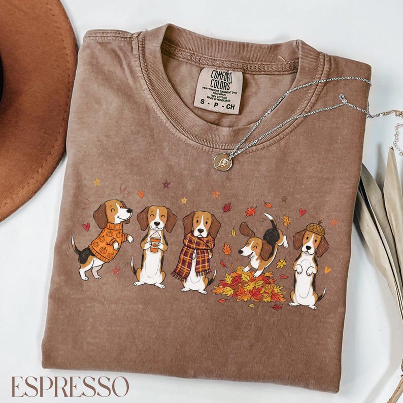 Puede incluir: Una camiseta marr&oacute;n de manga corta con etiqueta Comfort Colors que presenta un gr&aacute;fico de cinco perros beagle con ropa de oto&ntilde;o, incluyendo su&eacute;teres, bufandas y un sombrero de bellota, rodeados de hojas que caen y estrellas peque&ntilde;as.