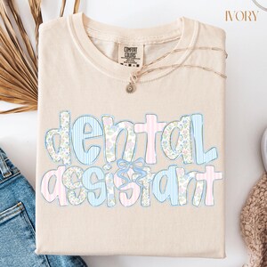 Op de afbeelding: Ivoor kleurig T-shirt met de woorden "dental assistant" in een speels lettertype. De tekst heeft een mix van bloemen- en streepmotieven in pastelblauw en roze, met een blauwe strik accent. Een gouden ketting is over het shirt gedrapeerd.