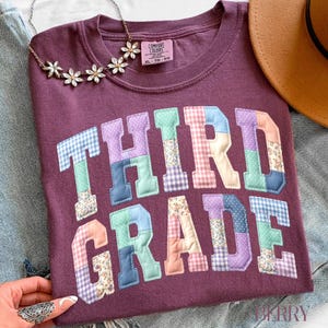 May include: A maroon Comfort Colors t-shirt with the words "THIRD GRADE" appliqued in colorful patchwork fabric. The shirt is size XL-TG and is accessorized with a floral necklace and a brown hat.