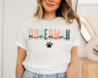 May include: White t-shirt with the text "DOBERMAN mama" in pastel colors and a black paw print. The shirt has rolled-up sleeves and a relaxed fit, perfect for casual wear.