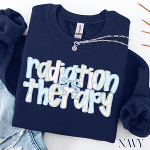 May include: Navy blue sweatshirt with the words "radiation therapy" in a floral and striped design. A silver necklace with a small pendant is draped across the sweatshirt. The word "NAVY" is at the bottom right.