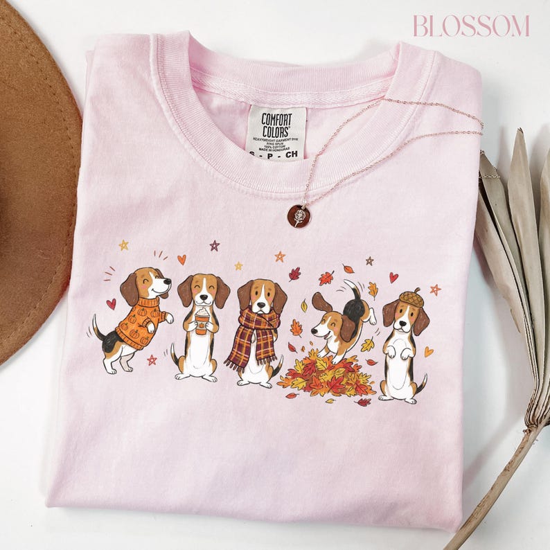 Puede incluir: Una camiseta de manga corta de color rosa claro con un gr&aacute;fico de cinco perros beagle con ropa de oto&ntilde;o, incluyendo jers&eacute;is, bufandas y un sombrero de bellota, rodeados de hojas que caen, corazones y estrellas. Tiene una etiqueta de marca.