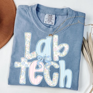 May include: A blue Comfort Colors t-shirt with the words "Lab Tech" in a floral and striped design. The shirt has a small bow detail and a gold necklace. The shirt is folded and has a small tag.
