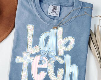 May include: A blue Comfort Colors t-shirt with the words "Lab Tech" in a floral and striped design. The shirt has a small bow detail and a gold necklace. The shirt is folded and has a small tag.