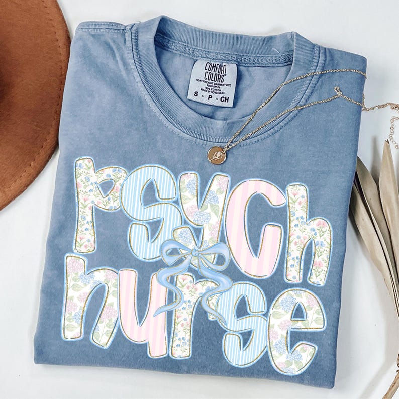 May include: A blue Comfort Colors t-shirt featuring the words "Psych Nurse" in a floral and striped design. A gold necklace with a pendant rests on the shirt. The t-shirt is neatly folded.