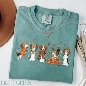 Puede incluir: Una camiseta verde claro con un gr&aacute;fico de cinco perros beagle con ropa de oto&ntilde;o, incluyendo jers&eacute;is, bufandas y un sombrero de bellota, rodeados de hojas ca&iacute;das. La camiseta tiene una etiqueta de la marca Comfort Colors en el cuello.