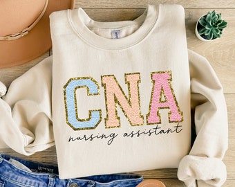 May include: A beige sweatshirt with the text "CNA nursing assistant" in gold lettering. The letters CNA are in a large font and are outlined in blue, pink, and peach.