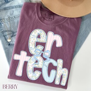 May include: A berry-colored t-shirt with the words "er tech" in a floral and striped design. The shirt is folded on top of a pair of blue jeans and a tan hat. The word "BERRY" is printed at the bottom left.