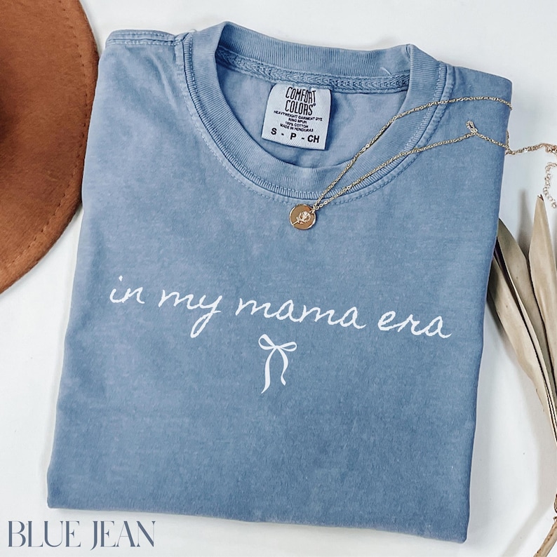 May include: A blue jean t-shirt with the text "in my mama era" in white script with a white bow underneath. The shirt has a distressed look and is made by Comfort Colors.