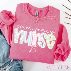 Puede incluir: Una sudadera rosa con las palabras "emergency nurse" en una fuente caprichosa. Las letras est&aacute;n decoradas con patrones florales, a rayas y de cuadros. Un delicado collar con un colgante descansa sobre la camisa. El texto "SAFETY PINK" est&aacute; en la parte inferior.