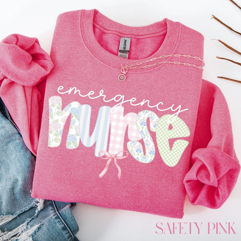 Puede incluir: Una sudadera rosa con las palabras "emergency nurse" en una fuente caprichosa. Las letras est&aacute;n decoradas con patrones florales, a rayas y de cuadros. Un delicado collar con un colgante descansa sobre la camisa. El texto "SAFETY PINK" est&aacute; en la parte inferior.