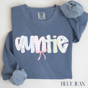 May include: A blue jean colored sweatshirt with the word "auntie" in a decorative font. The letters are patterned with stripes, gingham, and floral designs. A pink bow is centered below the word. The text "BLUE JEAN" is at the bottom.