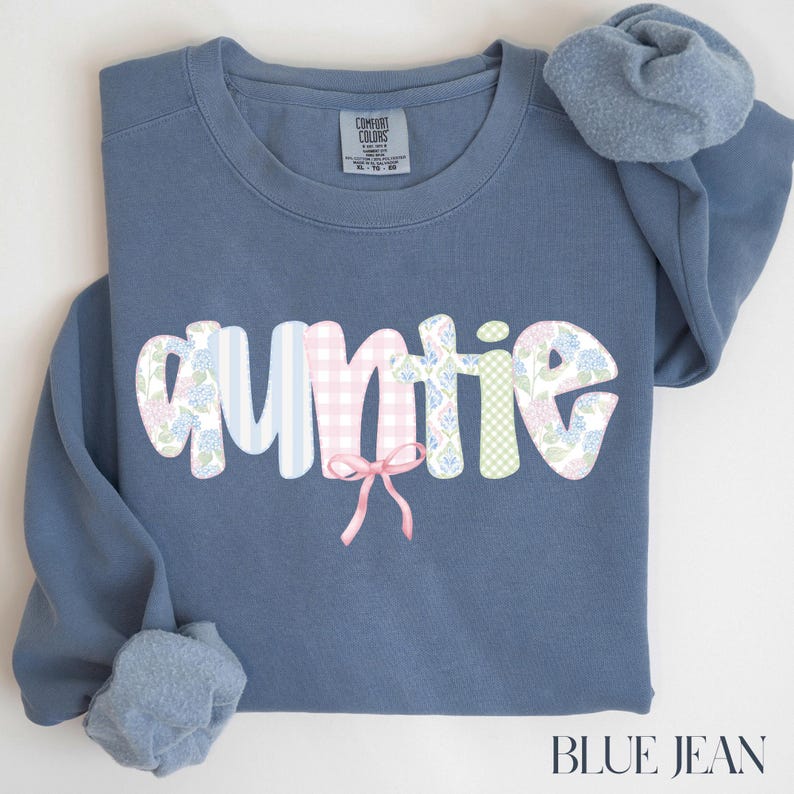 May include: A blue jean colored sweatshirt with the word "auntie" in a decorative font. The letters are patterned with stripes, gingham, and floral designs. A pink bow is centered below the word. The text "BLUE JEAN" is at the bottom.